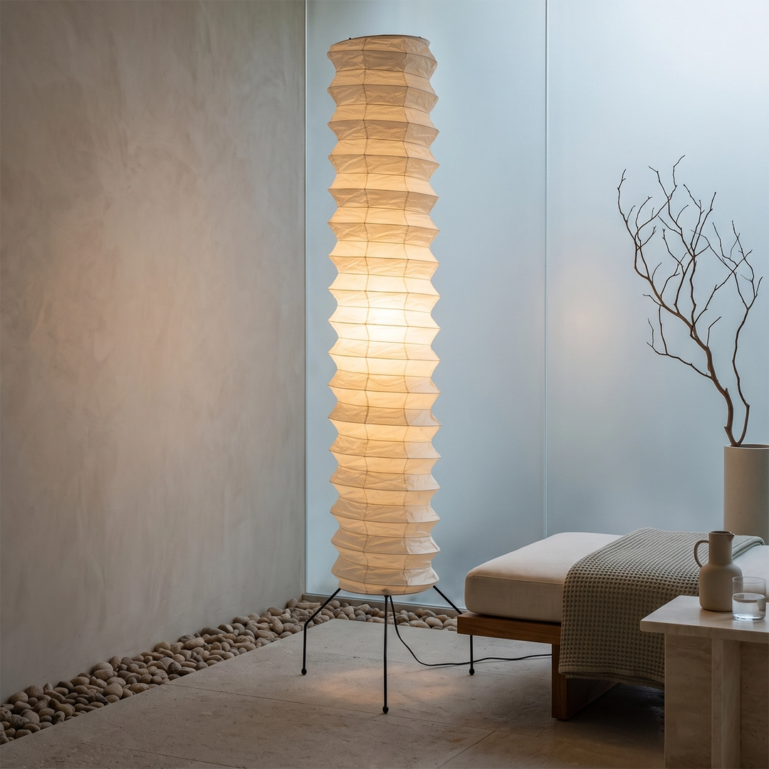 Cylinder paper floor lamp glowing in a corner against a soft blue wall beside bare branches