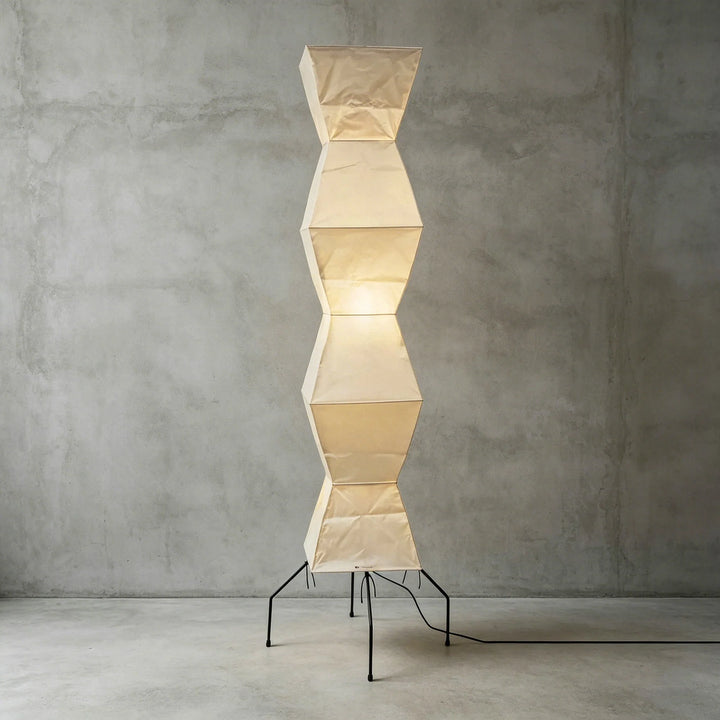 Faceted paper floor lamp lit against a gray concrete backdrop showing its stacked geometric form