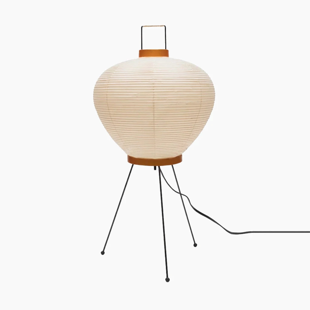 Rounded lantern paper table lamp with a black wire carry handle on top, an amber collar ring at the base and slim black tripod legs, shown unlit on a white background