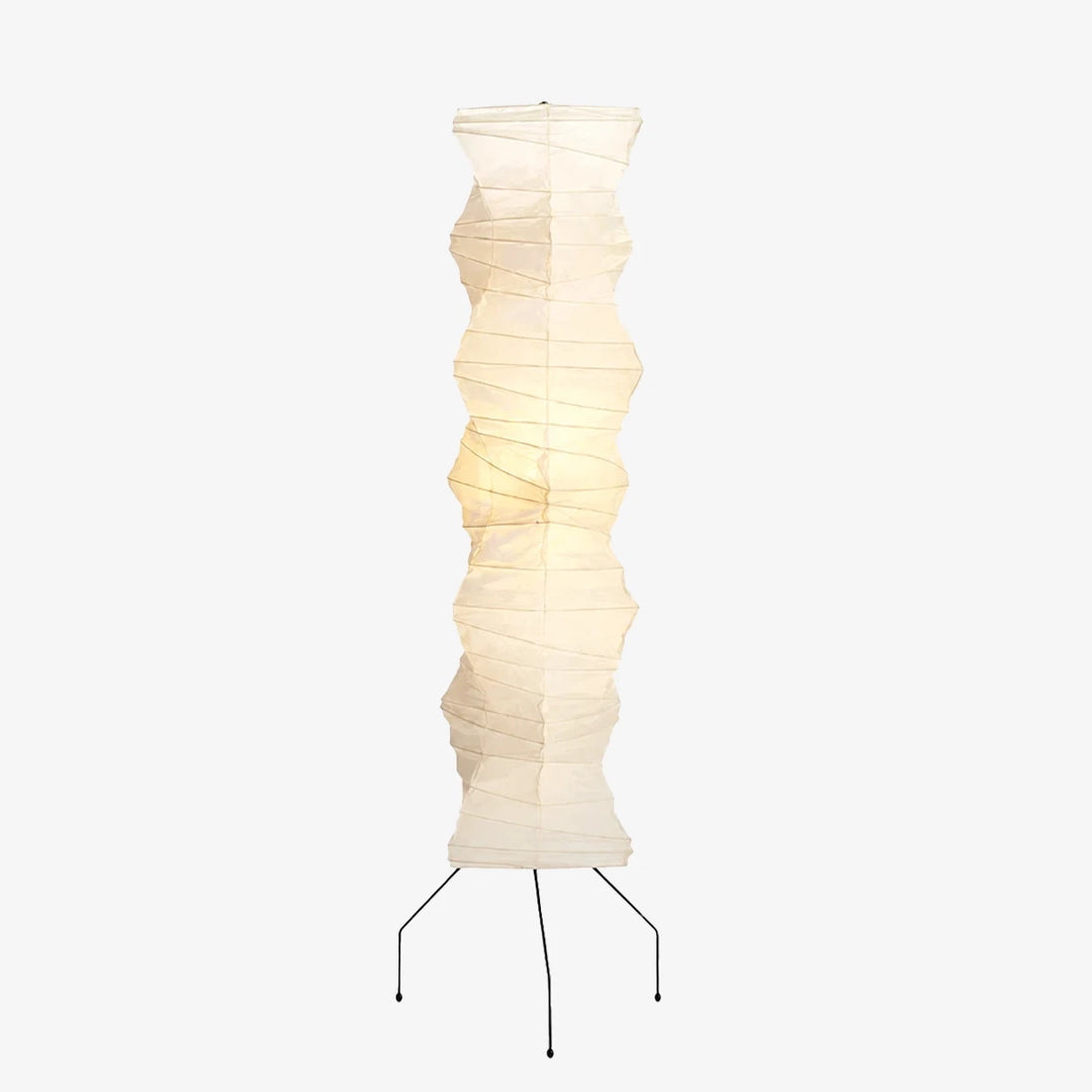 Tall sculptural faceted paper column floor lamp glowing warm with a tripod base on a soft grey background