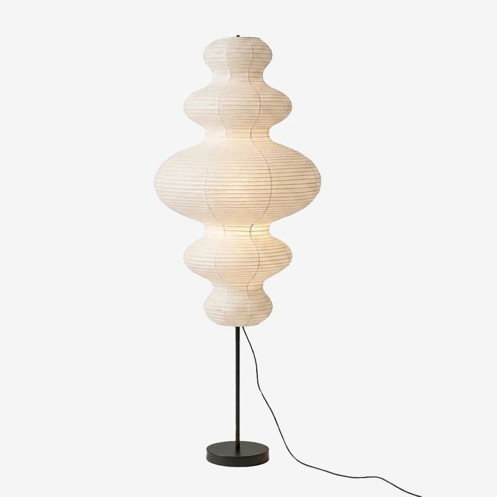 Sculptural paper floor lamp with stacked curved segments glowing softly above a slim black stem and round base, shown on a light gray background