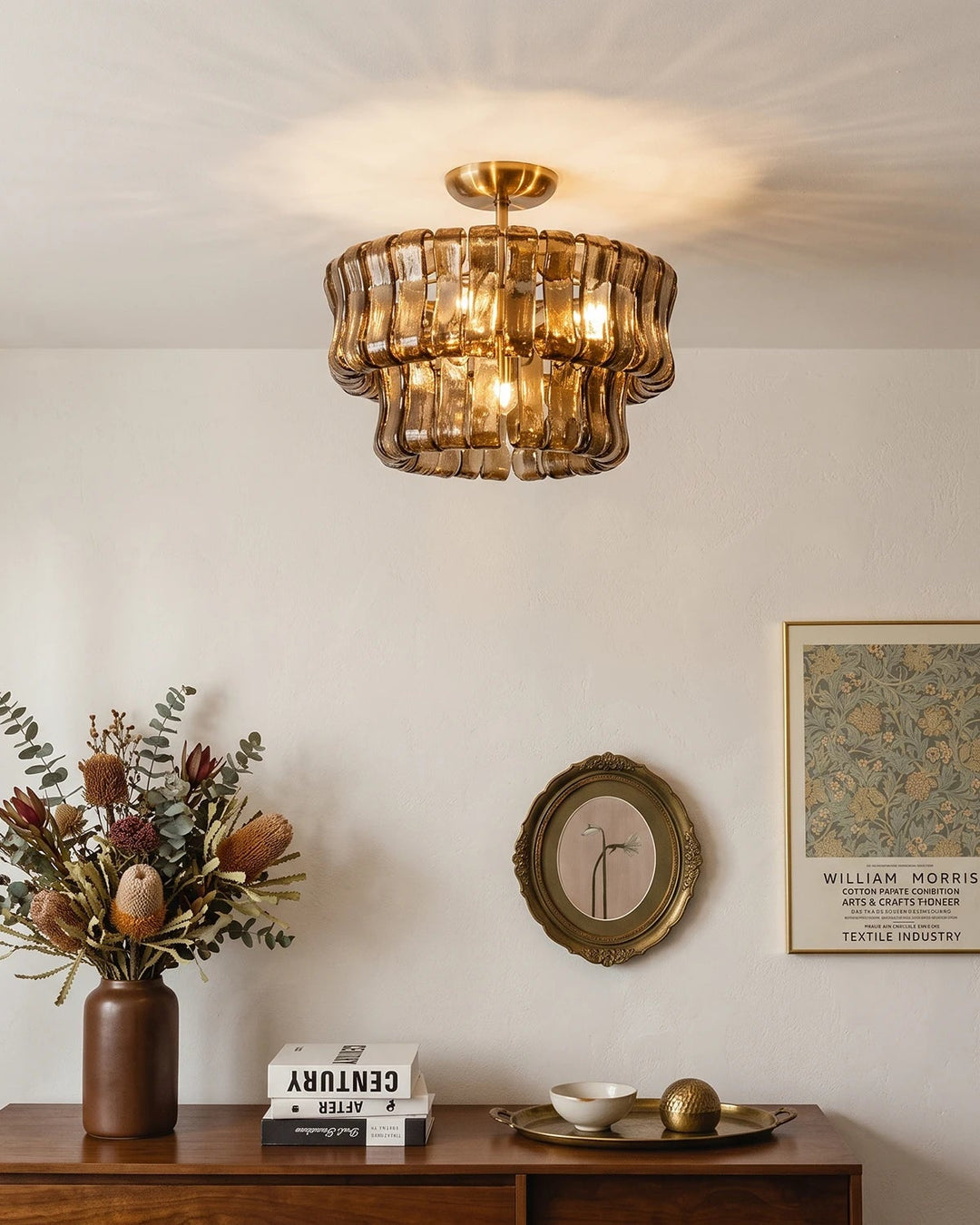 Smoke glass semi flush mount light lit above a wooden sideboard with flowers and framed art