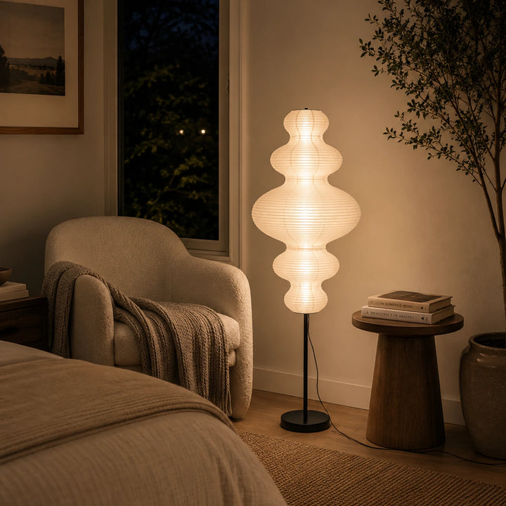 Stacked paper floor lamp casting a warm glow beside a bed at night