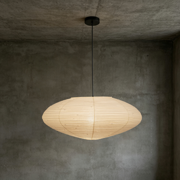 Warm glowing oval washi paper pendant lamp with black cord hanging on rough concrete wall